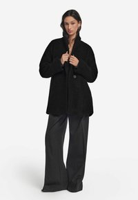 Czarny puszysty płaszcz z dużym kołnierzem i pojedynczym zapięciem na guzik, w połączeniu z szarymi spodniami o szerokich nogawkach, tworzący warstwowy strój.