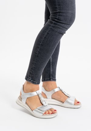 Silver metallic sandals with a textured upper, featuring an adjustable strap and prominent buckle detail, paired with dark skinny jeans.