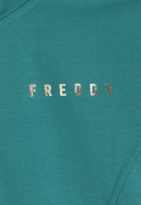 Il tessuto turchese presenta la scritta "FREDDY" in lettere metalliche argento. Il materiale è liscio con una leggera elasticità e un collo arrotondato.