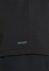 Camiseta deportiva negra con tejido que absorbe la humedad. Incluye el logo "NB DRY" impreso en azul claro cerca del dobladillo. Textura suave.