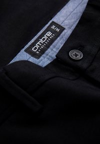 Pantaloni neri in tessuto con una cintura dotata di una fodera interna blu. Un piccolo bottone e un'etichetta con "Ombre Quintessence" sono visibili.