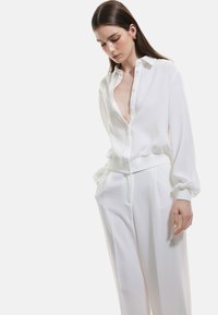 Blusa bianca con bottone frontale e colletto, dotata di maniche lunghe e vita elastica, abbinata a pantaloni bianchi a vestibilità larga coordinati.