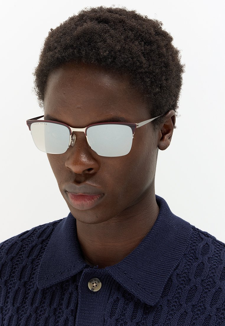 Braune, reflektierende Sonnenbrille mit einem schmalen, kastanienbraunen Rahmen und silbernen Akzenten. Das Modell trägt ein navyfarbenes, strukturiertes Strickpoloshirt.