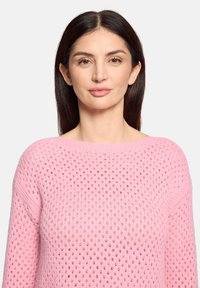 Femme aux cheveux raides et foncés portant un pull en tricot rose clair avec un motif texturé, tournée vers l'avant avec une expression neutre.