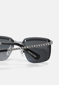Melnas saulesbrilles ar pelēkiem stikliem, embellētas ar rindu caurspīdīgu rhinestone pa tempļiem. Uz rokas ir redzams logo.