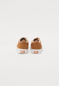 Baskets basses en suede marron avec des semelles en caoutchouc blanc et des accents orange, arborant le logo classique de Vans et des détails contrastants au niveau du talon.