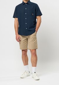 Marinblå kortärmad skjorta med knappslå, bröstficka och logotyp. Matchad med beigefärgade shorts och vita sneakers med strumpor.