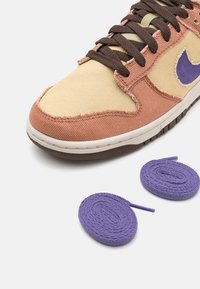 Sneaker i gul kordfløyel och rosa tyg med lila detaljer; har bruna snör. Två runda lila snör finns placerade bredvid.