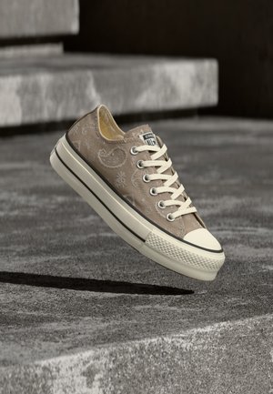 Beżowy niski sneaker z wzorem paisley, białymi sznurówkami i grubą białą podeszwą unoszącą się nad betonowymi schodami.