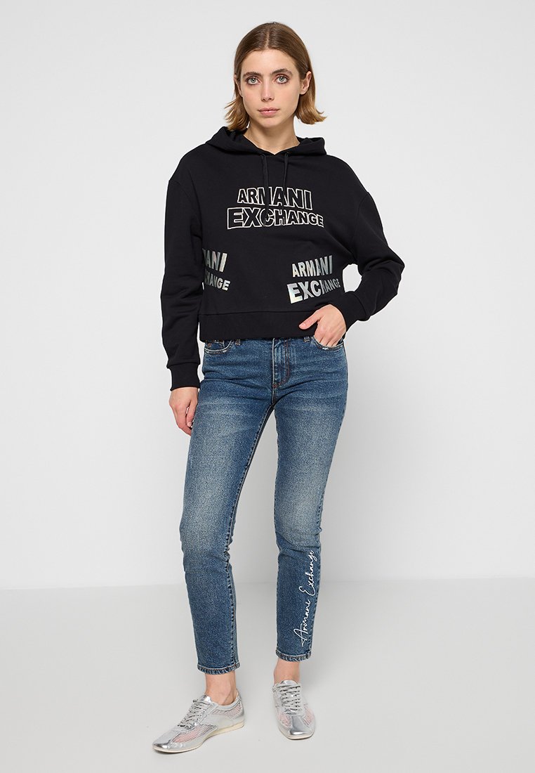Sudadera negra recortada con el logo de 'Armani Exchange' impreso en blanco, combinada con jeans azules. El modelo también lleva puestas zapatillas deportivas plateadas.