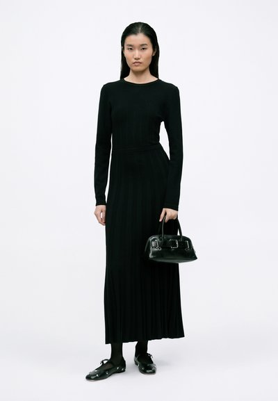 Vestido preto de malha canelada com mangas longas, apresentando um corte justo, uma cintura ajustada e uma bainha até ao chão. A modelo segura uma pequena bolsa preta.