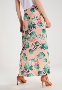 Žena v broskvovém maxi sukni se zeleným tropickým listovým a růžovým květinovým potiskem, spárovaná s korálovými vysokými podpatky, stojí bokem.