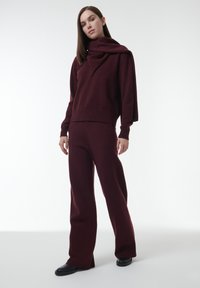 Bordeaux gebreid sweater met een vastgezette gedrapeerde sjaal en bijpassende flared broek. Zachte textuur, geribbelde manchetten en een ontspannen pasvorm. Zwarte schoenen.