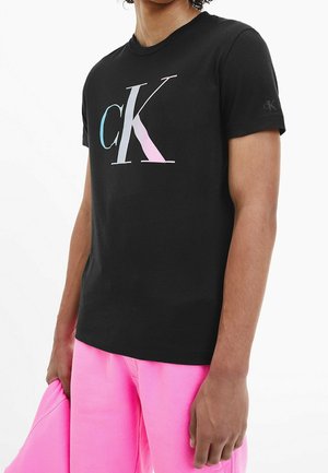 Zwarte katoenen t-shirt met een groot gradiënt "CK" logo. Korte mouwen, ronde hals, zichtbare branding op de mouw. G gedragen met roze broek.