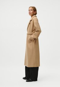Cappotto lungo beige con cintura, maniche ampie e tasche laterali, realizzato in tessuto morbido. Abbinato a pantaloni neri a gamba larga e scarpe nere.