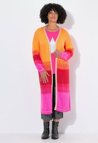 Donna con capelli ricci che indossa un lungo cardigan a righe arancione, rosso e rosa, una maglietta rosa brillante con una stella, pantaloni grigi e stivali neri con borchie.