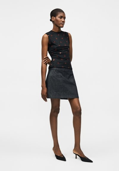 Γυναίκα στέκεται με το ένα χέρι κρατώντας τον αντίθετο αγκώνα, φοράει μαύρο αμάνικο τοπ με κόκκινα λουλουδάτα σχέδια, σκούρα φούστα και μαύρα τακούνια.