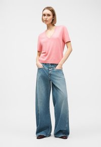 Femme aux cheveux blonds courts portant un haut rose à manches courtes avec un col en V et un jean large fluide bleu, les mains dans les poches.