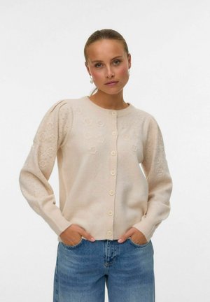 Femme avec des cheveux châtain clair en queue de cheval basse, portant des boucles d'oreilles en perles, un cardigan crème à boutons avec des motifs floraux en tricot, et un jeans bleu.
