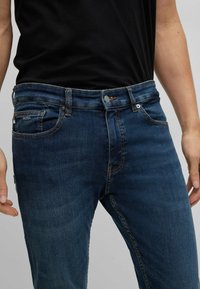 Hombre vestido con jeans azul oscuro con costuras naranjas y una camiseta negra, de pie contra un fondo neutro.