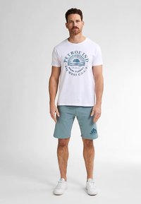 Witte katoenen t-shirt met blauwe grafische print, gecombineerd met lichtblauwe korte broek met logo. Model draagt witte sneakers.
