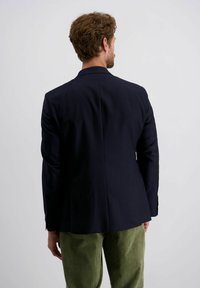 Man met krullend haar, gekleed in een donkerblauwe blazer en groene corduroy broek, met zijn rug naar de camera, tegen een lichte effen achtergrond.