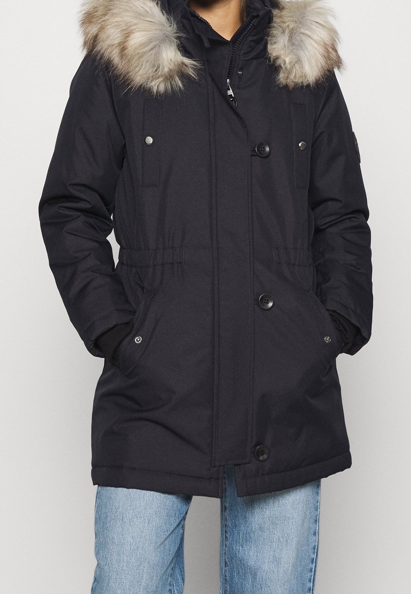 Marinblå parka med huva av fuskpäls, knappar och tryckknappar, elastisk midja och två framfickor. Har en vanlig passform och långa ärmar.