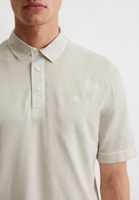 Jasno beżowa koszulka polo wykonana z teksturowanej tkaniny. Posiada klasyczny kołnierzyk, listwę z jasnymi guzikami oraz haftowane logo na piersi.