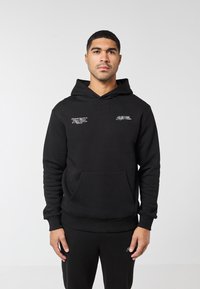 Mann trägt eine schwarze Kapuzenjacke mit weißem Text auf der Brust und schwarze Hose, steht vor einem einfarbigen hellgrauen Hintergrund.