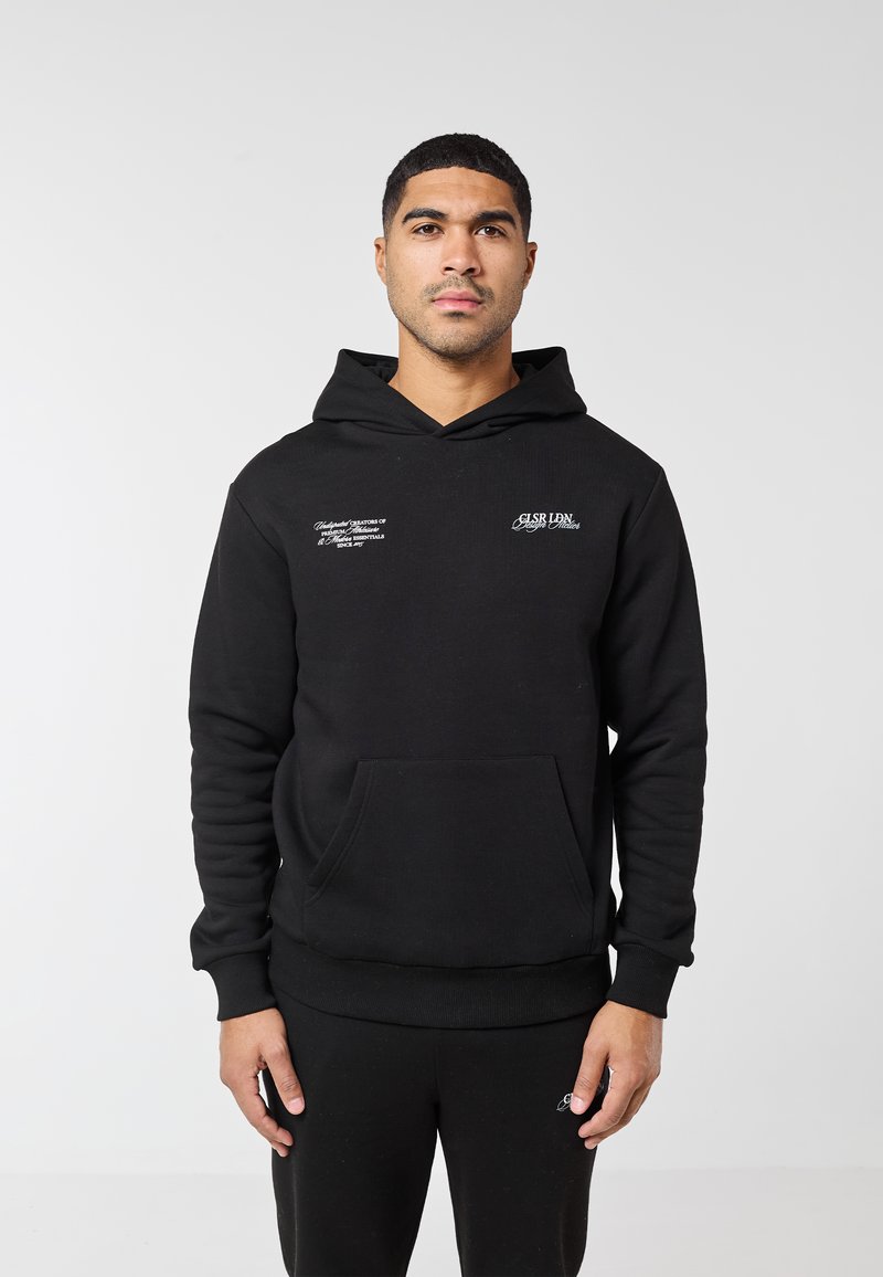 Mann trägt eine schwarze Kapuzenjacke mit weißem Text auf der Brust und schwarze Hose, steht vor einem einfarbigen hellgrauen Hintergrund.