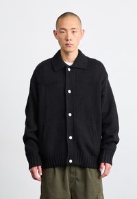 Cardigan lavorato a maglia nero con colletto e orlo a costine, dotato di bottoni bianchi e tasche laterali. Texture spessa e vestibilità morbida.