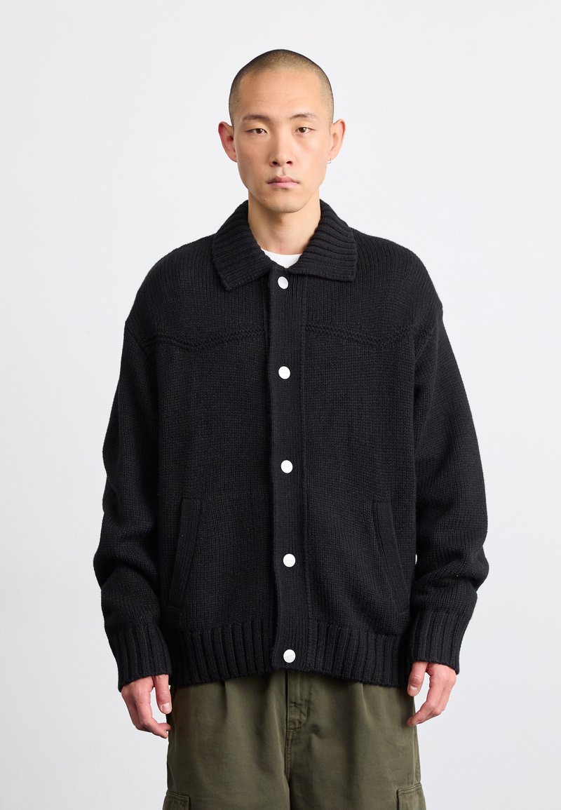 Cardigan lavorato a maglia nero con colletto e orlo a costine, dotato di bottoni bianchi e tasche laterali. Texture spessa e vestibilità morbida.