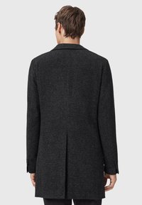 Svart texturerad kappa med rak passform, enkel slit bak och slagkrage. Utrustad med knäppte manschetter och ett subtilt vävt mönster.