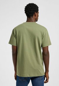 T-shirt in cotone verde oliva con vestibilità ampia, colletto rotondo e maniche corte, mostrata da dietro su uno sfondo neutro.