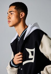 Zwarte en crèmekleurige varsityjack met geborduurd logo, grijze capuchon en gestreepte accenten. Model afgebeeld met hand op de jasopening.