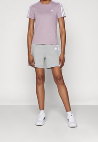 Svetle fialové tričko s krátkým rukávem a bílým designem se třemi pruhy a logem, spárované se šedými šortkami s logem a pruhy, a bílými sportovními botami.