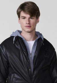 Giubbotto bomber nero in nylon con cuciture a contrasto, chiusura con zip e una felpa grigia sotto. Presenta una "K" ricamata sul petto.