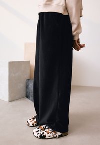 Crne hlače širokih nogavica od mekane tkanine, uparene s smeđim i bijelim papučama s uzorkom. Ruke su opuštene uz tijelo, na neutralnoj pozadini.