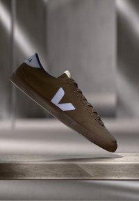 Smeđa tenisica od antilopa s bijelim logotipom i plavim detaljem na peti. Ima teksturiranu gumenu potplatu i klasični dizajn s vezicama.