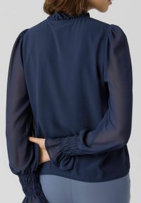 Blouse bleu marine avec manches longues transparentes, poignets froncés et col montant. Texture lisse avec une coupe décontractée et des détails subtils aux épaules.