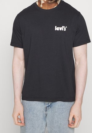 Osoba w czarnej koszulce Levi’s z białym logo na piersi, w połączeniu z jasnoniebieskimi dżinsami, stoi na jednolitym tle.