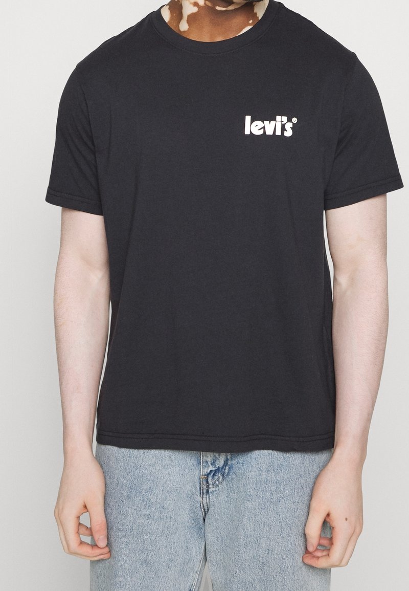 Personne portant un t-shirt Levi's noir avec un logo blanc sur la poitrine, assorti à un jean bleu clair, debout devant un fond uni.
