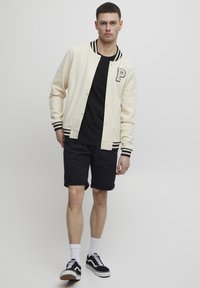 Cream varsity jacket with black stripes, black t-shirt, dark shorts, and black sneakers. Features a prominent "P" on the chest.