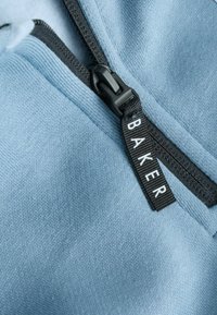 Hellblauer Stoff mit einer strukturierten Oberfläche, versehen mit einem schwarzen Reißverschluss und einem Etikett mit der Aufschrift "BAKER". Das Design ist schlicht und funktional.