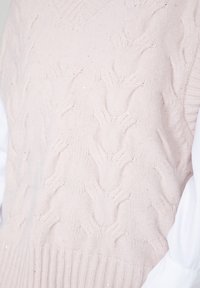 Lichtroze gebreide sweatervest met een gestructureerd kabelpatroon, V-hals ontwerp en ribbel onderkant. De achtergrond is gedeeltelijk onduidelijk.