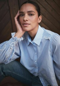 Kvinde med mørkt hår sat tilbage, hvilende hovedet på hånden, iført en blåstripet bluse med blonde detaljer og mørke jeans, foran en træbaggrund.