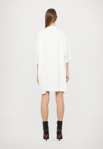 Fehér, oversize rövid ujjú ruha kerek nyakkivágással, amely laza szabású és minimalista dizájnú. Fekete bokacsizma piros kiegészítőkkel.