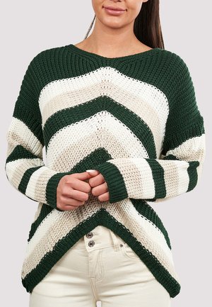 Kvinde iført en grøn og hvid stribet strikket sweater med chevronmønster, stående med hænderne foldet foran, kombineret med lyse bukser.