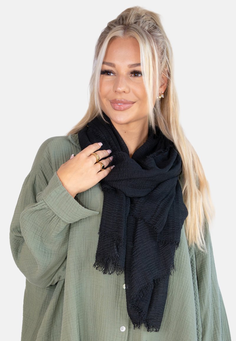 Écharpe noire texturée avec des bords effilochés, portée autour du cou, associée à une chemise verte. Met en avant le matériau doux et le design superposé.