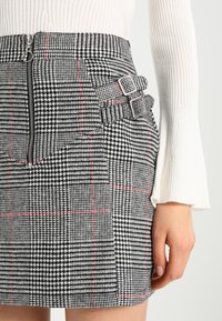 Mini gonna in bianco e nero con motivo a scacchi houndstooth, dotata di zip laterale, con linee rosse e dettagli a doppia fibbia in vita.